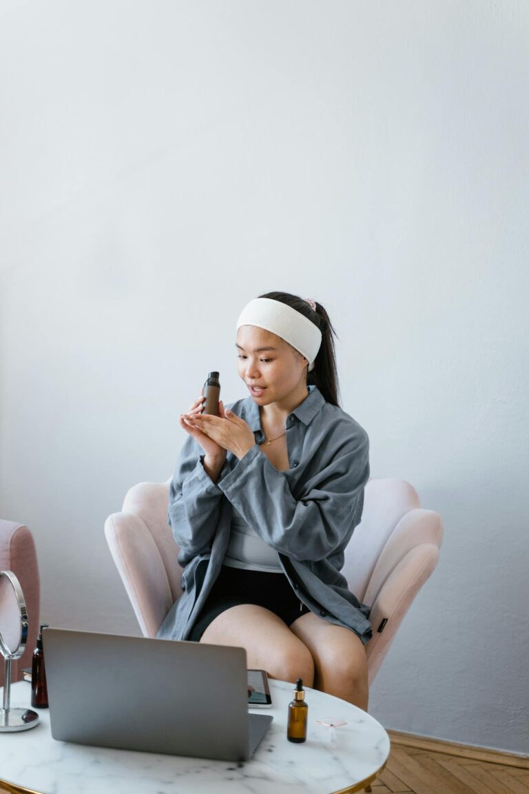 Young woman with headband reviews beauty products at home using laptop.